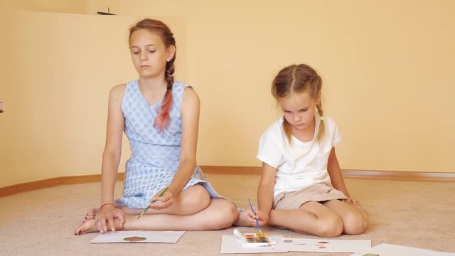 Charming sisters sitting on floor with papers and drawing with watercolors at home