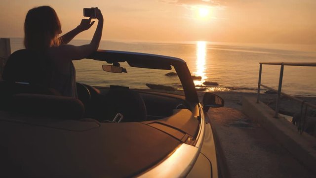 Woman in luxury car takes a photo of sunset over sea by her touchscreen cell phone
