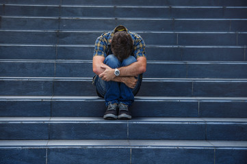 young sad and desperate man sitting outdoors at street stairs suffering anxiety and depression feeling miserable crying in unemployment and broken heart concept