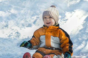 Boy in hat and orange jumpsuit slides off snow slide on back. Portrait. Close-up. Winter day.