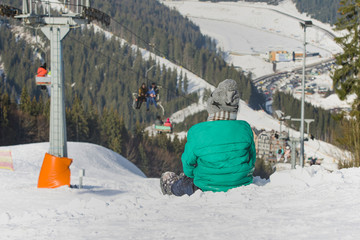 Boy sits on a snow-covered hill on background of cable car, pine forest and mountains. Ski resort. Winter day