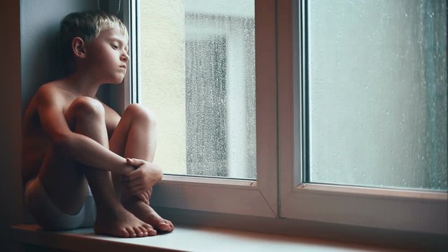 Sad boy siting on windowsill on a rainy day, looking out of window and thinking something sorrow. Little kids upbringing lifestyle concept video.