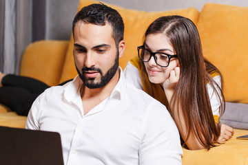 Pretty long-haired girl in pullover wearing glasses laying on the yellow sofa in the light living room and looking over shoulder how her bearded multicultural boyfriend working on the laptop, modern