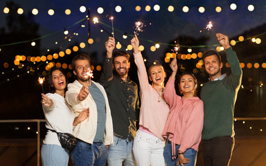 leisure, celebration and people concept - happy friends with sparklers at rooftop party at night...