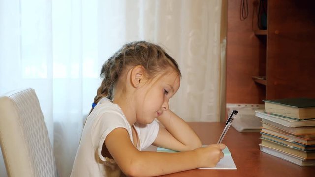 Little girl sitting at table with pile of books and doing homework leaning on hand