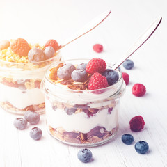 Two jars with tasty parfait made of granola, berries and yogurt on white wooden table. Shot at...