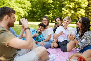 friendship, leisure and technology concept - man with camera photographing his friends drinking non alcoholic drinks at picnic in summer park