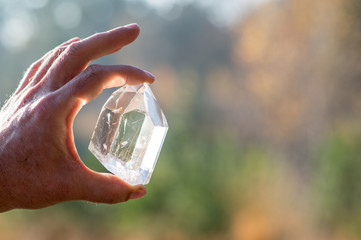 A Hand holding a transparent crystal quartz with the sun hitting the gemstone and a blurry background - obrazy, fototapety, plakaty