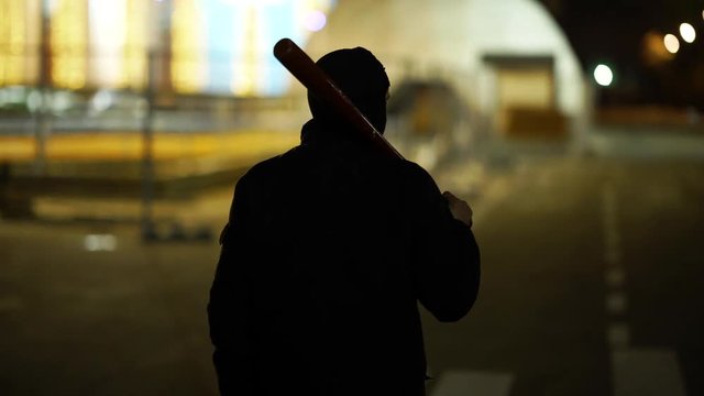 A man dressed in dark clothes and a balaclava goes with a bat in an abandoned park. There is an early winter outside and garlands hang around. His face is not visible in backlight. He is set up to fig