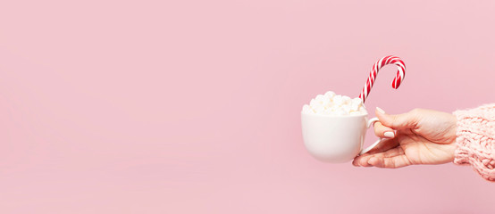 Female hands in knitted sweater holding cup of marshmallows and Christmas candy cane on pink background Flat Lay copy space. Winter traditional food Festive decor celebration presents Xmas holiday