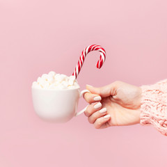 Female hands in knitted sweater holding cup of marshmallows and Christmas candy cane on pink background Flat Lay copy space. Winter traditional food Festive decor celebration presents Xmas holiday