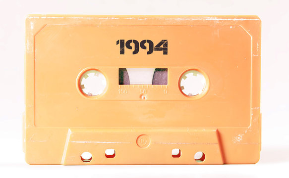A vintage cassette tape from the 1980s era (obsolete music technology) with the text 1994 printed over it, stencil font. Color: cream, sand. White background.
