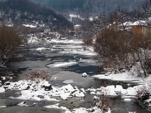 Picturesque elevated snowy rural view from a bridge to the winter Vidima River and old riverbank houses at the Bulgarian village of Debnevo, Central-North Bulgaria