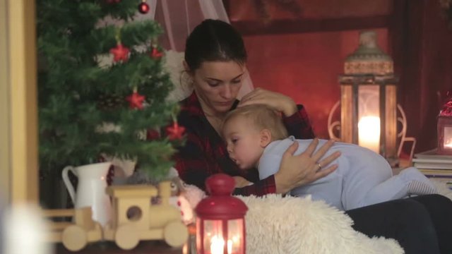 Mother breastfeeding her toddler son sitting in cozy armchair near Christmas tree, wintertime