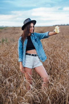 Young woman wearing hat and denim jacket taking selfie with camera in a corn field