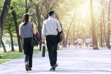 Fototapeta premium The back of couple businessman and businesswoman walking together within the park relaxed after work. Two businessmen holding cup of coffee walk talk happily in the park. Relaxing concept