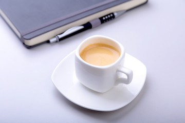 white cup with a fragrant espresso coffee, black stylish notebook and ballpoint pen on the office desk.