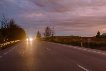 Vehicle with bright headlamps riding along asphalt road in amazing countryside in cloudy evening