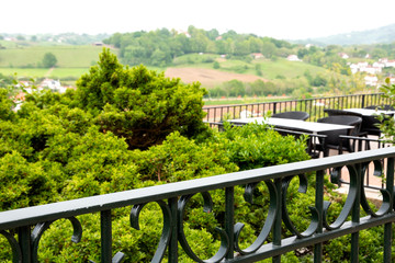 Fototapeta na wymiar French Basque country, Beautiful view of green hills and valley with village houses from Cambo-les-Bains (charming spa town) promenade. Empty cafe terrace tables. Springtime. France. Selective focus