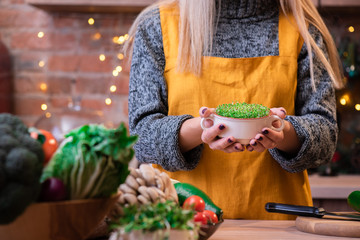 Girl with blond hair in a yellow apron in a loft kitchen, decorated with garlands, holds a bowl...