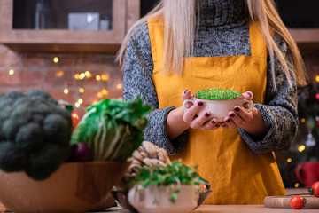 Girl with blond hair in a yellow apron in a loft kitchen, decorated with garlands, holds a bowl with microgreen/sprouts. Wooden plate with vegetables: broccoli, onions, cucumbers, microgreens etc