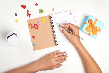 Female hands are writing on notebook. New Year 2019. Goals. Christmas planning concept . Gift box, decorations, empty notebook and coffee on white table. To do list. Mockup, copy space.