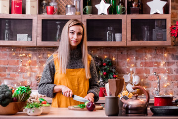 Young girl with blonde long hair and in a yellow apron cuts cucumbers in a bright loft kitchen. Assorted vegetables in a wooden plate on a light wooden table. close up