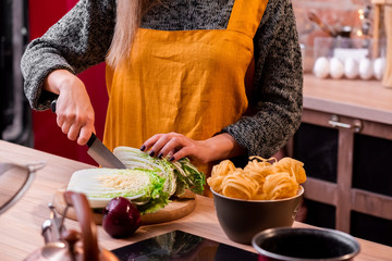 Smiling happy young girl of European appearance with blond haire  in a yellow apron at the loft kitchen is cuts Chinese cabbage. Variety of vegetables in a wooden plate on a light wooden table.