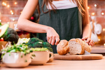 Girl in a green apron cuts the bread in a beautiful bright loft kitchen decorated with a garland....
