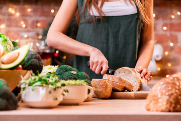 Girl in a green apron cuts the bread in a beautiful bright loft kitchen decorated with a garland. Assorted vegetables and fruits in a wooden plate on a light wooden table.