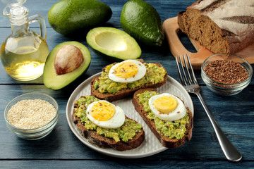 Sandwich with avocado puree and egg on a plate and ingredients for cooking on a blue wooden table.