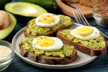 Sandwich with avocado puree and egg on a plate and ingredients for cooking on a blue wooden table.