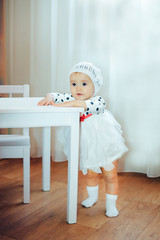 A little girl stands near a white table in a knitted hat