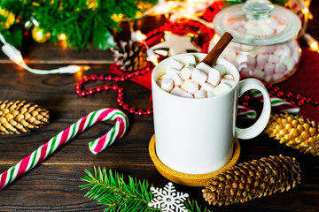 White mug of cocoa with marshmallows, lollipops, fir cones, Christmas tree branch, garland and snowflake on wooden table
