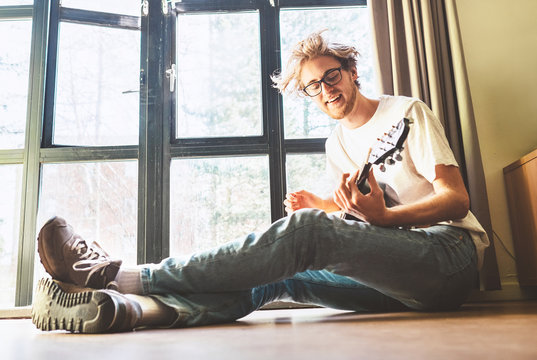 Young blondhair handsom man plays on guitar sitting on floor near the whole wall window at home. Music education concept image.