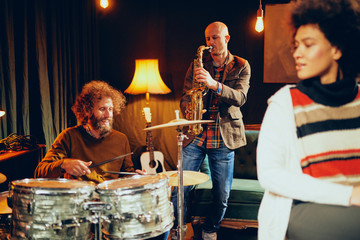 Multicultural band practicing for the gig in home studio. In foreground mixed race singer and in background drummer and saxophonist.