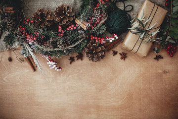 Stylish rustic gift box with green branch and wreath, fir,anise, pine cones,berries, cinnamon on rustic wooden background.  Space for text. Merry Christmas concept, atmospheric flat lay.