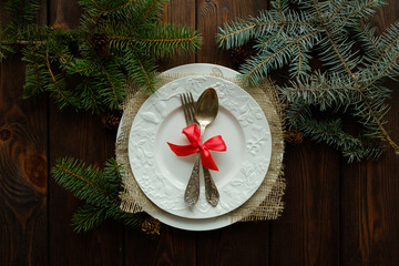 Antique or rustic table of Christmas tables from above. Elegant empty white plate, cutlery, red bow and natural pine on a wooden table.