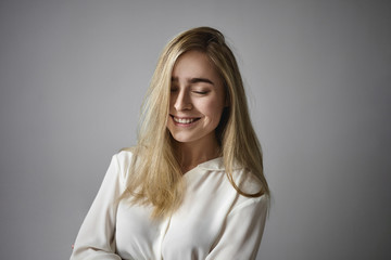 Positive human emotions, feelings, reaction and life perception. Picture of adorable cheerful young Caucasian lady with long fair hair keeping eyes closed and smiling broadly, being in good mood