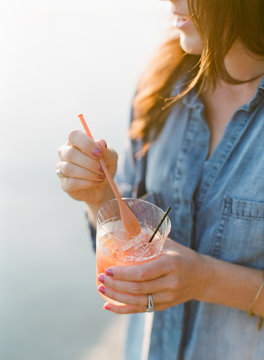 woman in denim shirt holding cocktail by the lake at sunset outside