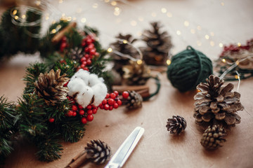 Fir branches, wreath, red berries, pine cones, thread, scissors, cinnamon, cotton, lights on rustic wooden background. Details for making christmas wreath at workshop. Atmospheric image