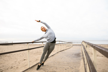 Full length portrait of fashionable flexible young sportswoman wearing hoodie, leggins and running shoes posing on wooden pier, doing yoga or stretching, having concentrated facial expression