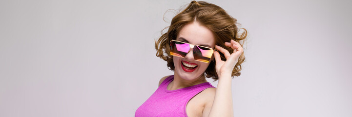 Naklejka premium Charming young girl in pink dress on gray background. Cheerful girl in square glasses. The girl adjusts glasses
