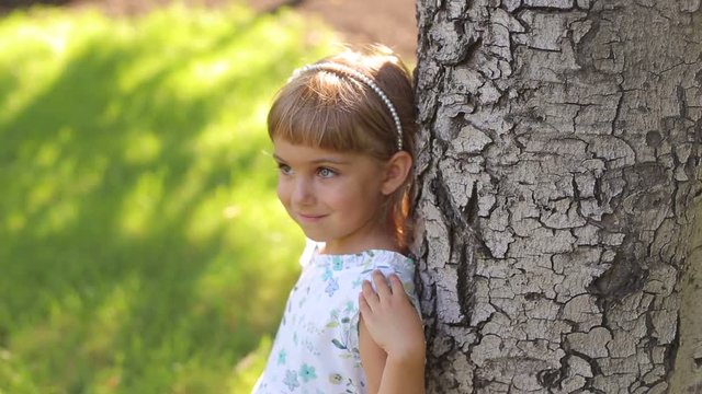 A little cute girl in a light summer dress with heterochromia stands leaning against a big tree in the Park. Portrait of a cute four-year-old girl with eyes of two different colors.