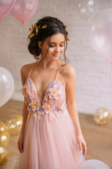 a gorgeous young queen with dark hair and gold in a chic light pink dress with purple color, confusedly lowers her eyes to the floor, stands in a bright spacious room near the balloons - obrazy, fototapety, plakaty