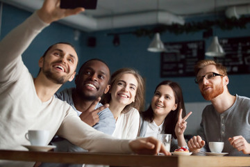 Excited millennial friends make selfie on smartphone having fun in coffeeshop, happy students smile for picture on phone meeting together in cafe, diverse young people posing for self-portrait