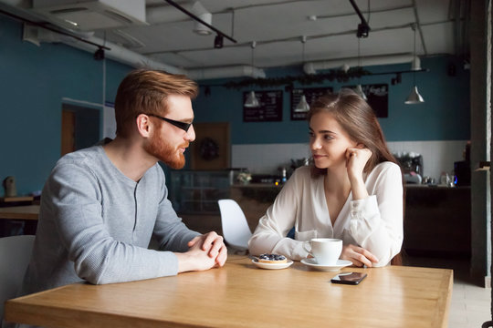 Millennial red-haired man approaching beautiful young girl enjoying coffee and dessert in cafe, interested young guy get acquainted with pretty female, young people flirt sitting in coffeeshop
