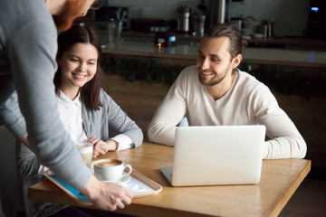 Waiter give order to smiling millennial couple sitting at coffeeshop table with laptop, young cafe worker bring cappuccino and latte to happy coffeehouse visitors, server serving cups with hot drinks