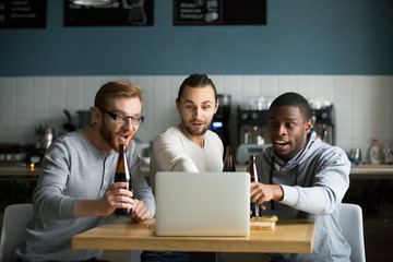 Diverse friends hang out in cafe drinking beer, watching online game at laptop, multiethnic males...