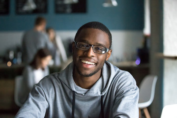 Portrait of smiling African American student looking at camera sitting in cafe, black millennial man posing making picture in coffeeshop, afro male in glasses drinking coffee working in coffeehouse - Powered by Adobe